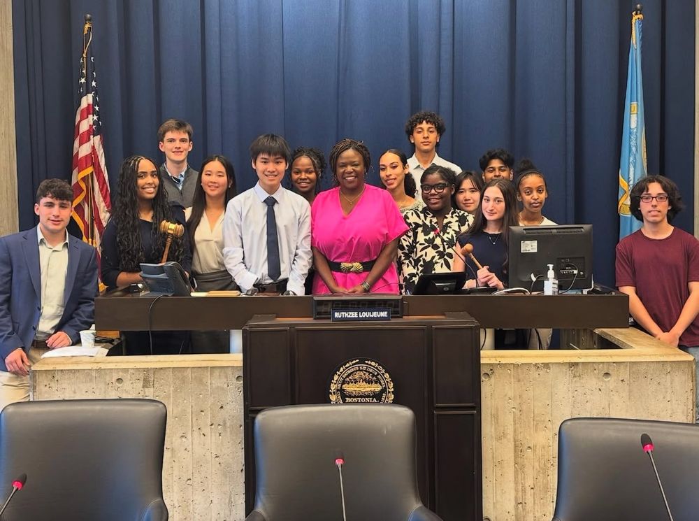 A group of approximately 15 young people, appearing to be high school or college students, pose together in what looks like a government meeting room or city hall chamber. They are gathered around and behind a speaker’s podium that has “RUTHZEE LOUIJEUNE” displayed on a nameplate. The person at the center of the group, wearing a bright pink outfit, appears to be Ruthzee Louijeune.
The students are dressed in a mix of business casual and formal attire, with some wearing suits and ties while others are in more casual clothing. Behind them are blue curtains, and both an American flag and what appears to be a state or municipal flag are visible on either side of the group. The setting has the formal appearance of a legislative chamber or city council meeting room, with tiered seating visible in the foreground. The students appear to be participating in some kind of civic engagement program, mock trial, or student government activity.​​​​​​​​​​​​​​​​