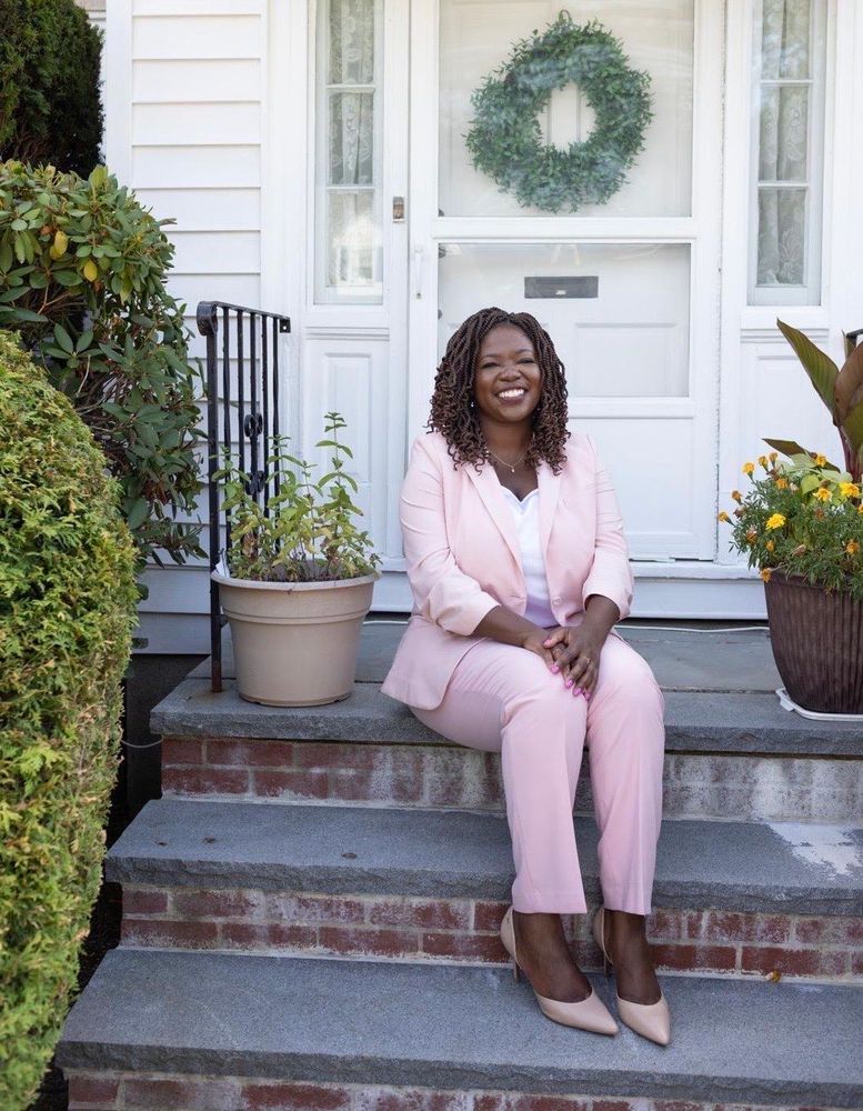 Ruthzee in a pink blazer and matching pants sits smiling on the front steps of a white house. She has shoulder-length braided hair and is wearing nude heels. The front porch features potted plants, a decorative wreath on the white door, and brick and stone steps. The setting is landscaped with various greenery and flowers visible around the entrance.​​​​​​​​​​​​​​​​