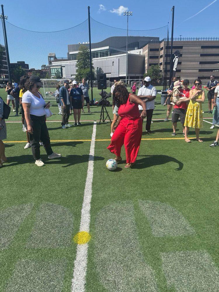 Ruthzee Louijeune in a red outfit demonstrates soccer skills on a turf field while spectators watch. She’s positioned near the center line with a soccer ball at her feet, surrounded by onlookers including someone in a white top and green pants. Modern buildings and stadium lighting are visible in the background.