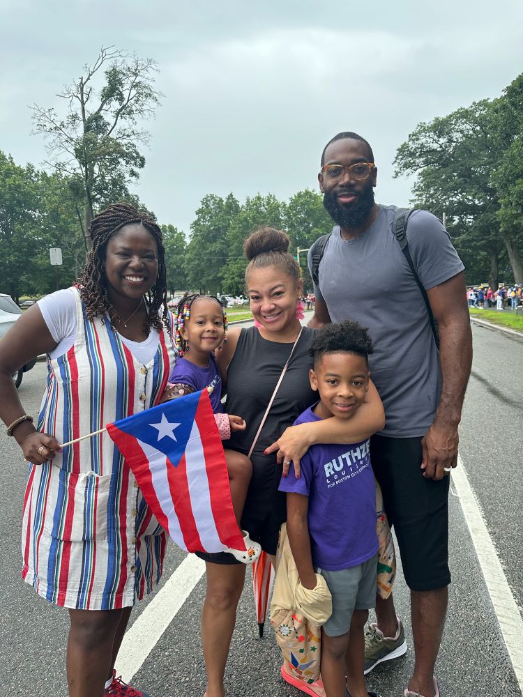 A smaller group photo on the same street showing Ruthzee Louijeune in her striped dress posing with a family of four, including two children. One person is holding a Puerto Rican flag. The backdrop shows the same tree-lined street with parked cars and other event attendees visible in the distance.