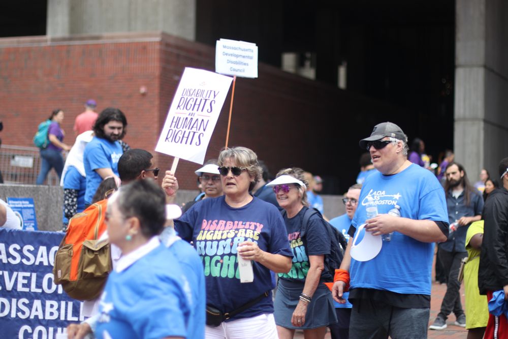 Protesters at a demonstration, with one person prominently holding a purple and white sign reading “DISABILITY RIGHTS ARE HUMAN RIGHTS.” Several attendees are wearing blue shirts, including one with “ADA 35 BOSTON” visible. There’s also a smaller sign mentioning “Massachusetts Developmental Disabilities Council.” The scene takes place near brick buildings in what appears to be a downtown area.​​​​​​​​​​​​​​​​