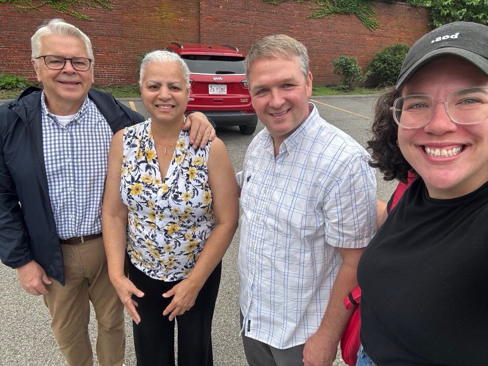 Four people posing together for a photo in a parking lot. From left to right: a man in glasses wearing a blue checkered shirt and dark jacket, a woman in a floral sleeveless top, a man in a light checkered shirt, and a woman in a cap taking a selfie. A red car and brick building are visible in the background.