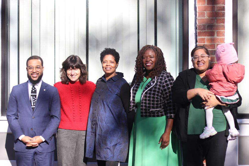 Five professionals standing in a line outside a modern building with white siding and brick accents. From left to right: a person in a navy suit, a person in a red sweater, a person in a navy coat, a person in a green dress with a black and white checkered blazer, and a person in a green dress holding a young child in pink clothing