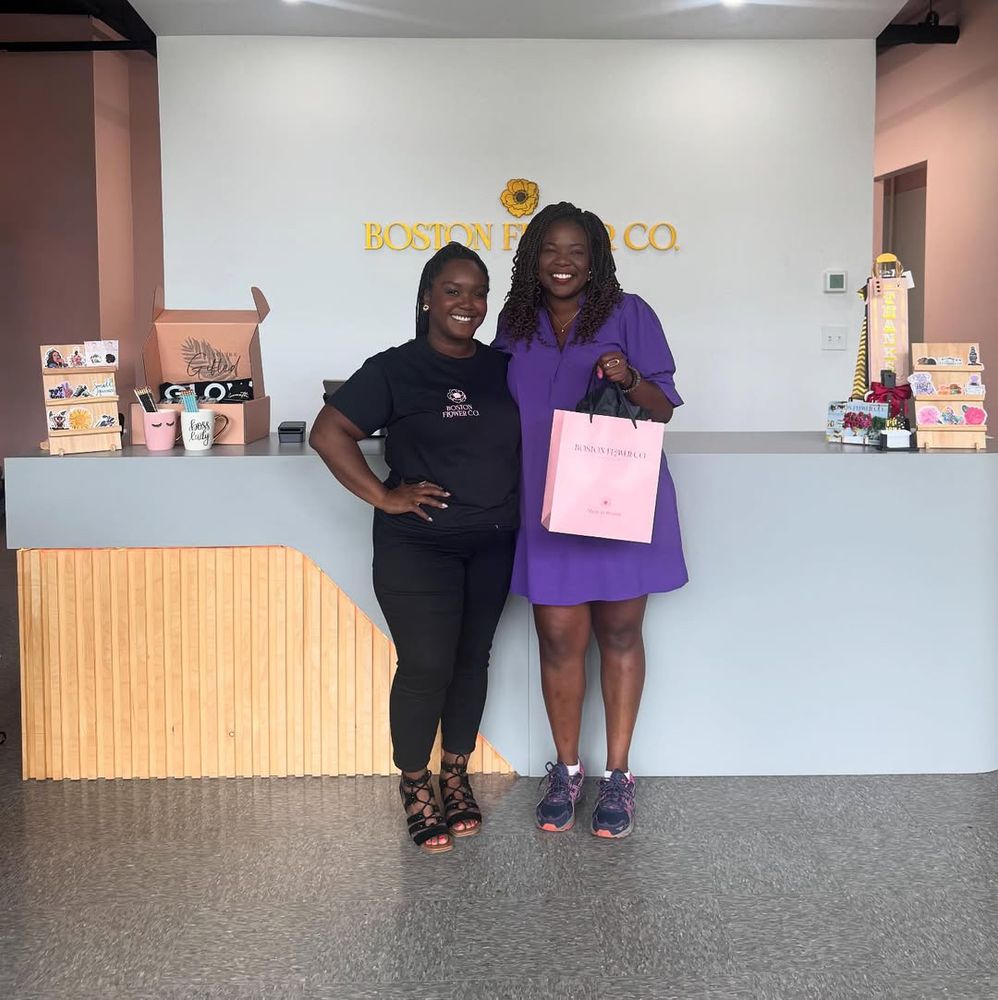Two smiling women posing together inside Boston Flower Co. store. One woman wears a black Boston Flower Co. t-shirt and black pants, while Ruthzee Louijeune wears a purple dress and holds a pink shopping bag. They’re standing in front of a white counter with the golden Boston Flower Co. logo on the wall behind them. The store interior features pink walls, wooden counter details, and various products displayed on shelves.