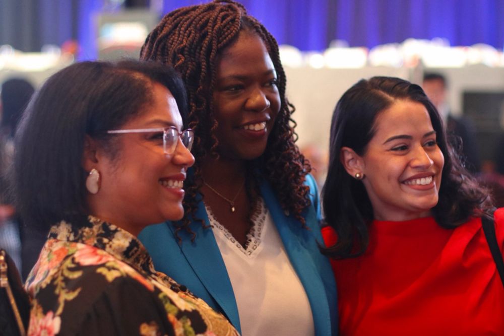 Three people are captured in a candid moment, all smiling and appearing to be at a formal event. The person on the left wears glasses and a floral print outfit, the person in the center, Ruthzee, wears a bright blue blazer, and the person on the right wears a red top. The background shows a blurred event space with blue lighting.