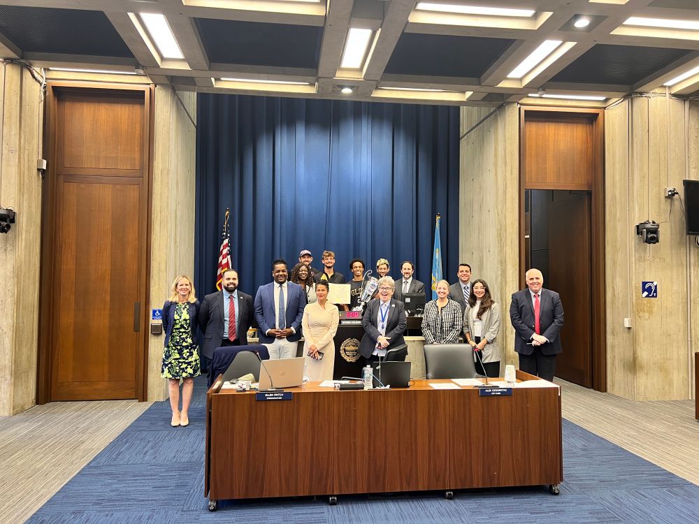 A large group photo taken in a municipal council chamber showing approximately 15-20 people gathered around and behind a wooden council desk. The chamber features a blue curtained backdrop, American flag, and modern architectural elements with a coffered ceiling. The group appears to be a mix of officials and community members, likely during a recognition ceremony or special council meeting.​​​​​​​​​​​​​​​​