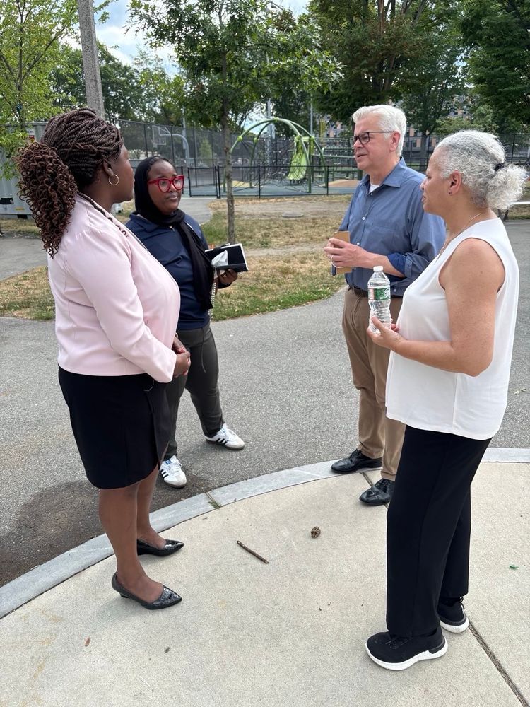 Four people having a conversation on a sidewalk near a park. Two women on the left, including Ruthzee Louijeune in pink blazer with braided hair and another in dark clothing with red glasses, are speaking with a man in a blue shirt and glasses and a woman in a white top on the right. Trees and playground equipment are visible in the background.