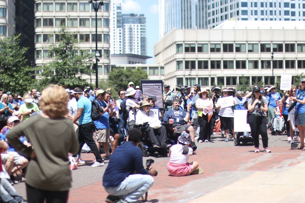A large crowd gathered in an urban plaza surrounded by office buildings and high-rises. Many attendees are wearing light blue shirts and some are seated in wheelchairs. People are holding various signs and banners, and the crowd appears to be participating in an organized demonstration or rally on a sunny day.
