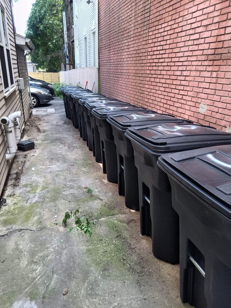 A narrow alleyway or side passage between buildings showing a long row of black wheeled garbage bins lined up against a red brick wall. The bins have dark lids and appear to be standard residential waste containers. The ground is concrete, and there are residential buildings with siding visible on the left side of the passage.​​​​​​​​​​​​​​​​