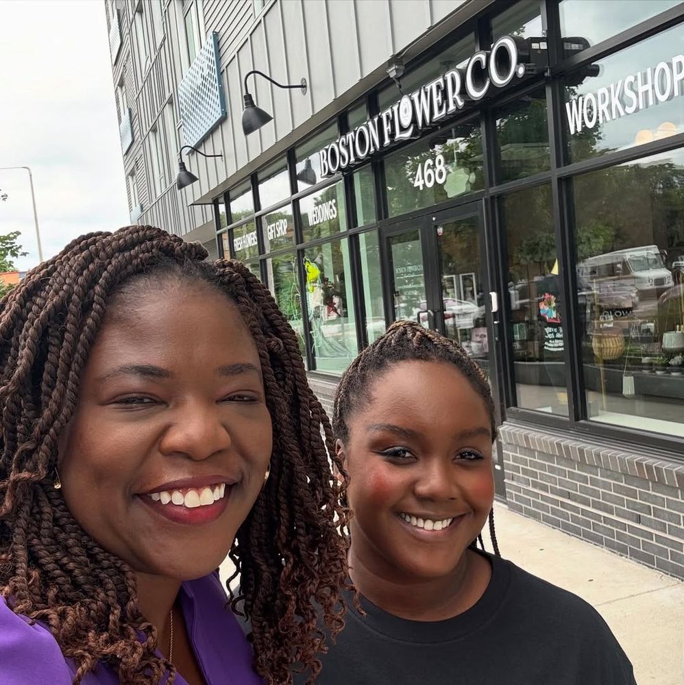 Two smiling women taking a selfie outside the Boston Flower Co. storefront at 468. Ruthzee Louijeune wears a purple top and has long twisted braids, while the other woman wears a black top and has shorter braided hair. Behind them is the store’s glass front with “BOSTON FLOWER CO.” and “WORKSHOP” signage visible, along with the address number 468.​​​​​​​​​​​​​​​​