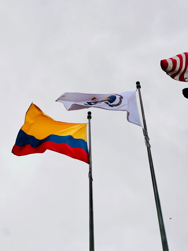 Three flags wave against an overcast sky - the Colombian flag with its distinctive yellow, blue, and red horizontal stripes is prominently featured on the left, alongside what appears to be a local government or organizational flag in the center, and a partial view of the American flag on the right.
These images appear to document a Colombian cultural celebration or official ceremony taking place at Boston City Hall Plaza.​​​​​​​​​​​​​​​​