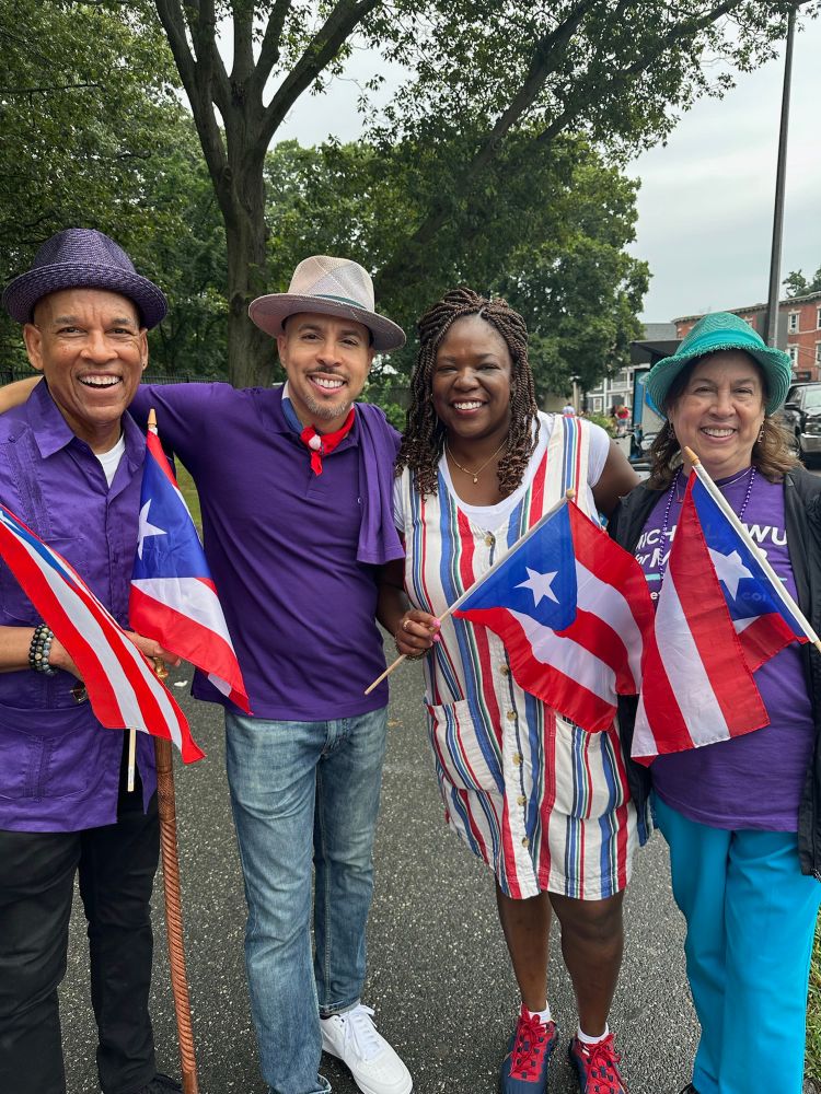 A group of four people wearing purple shirts and holding Puerto Rican flags pose together on the street. Ruthzee Louijeune in her striped dress stands third from left. Everyone is smiling and appears to be participating in the same community event or parade.​​​​​​​​​​​​​​​​