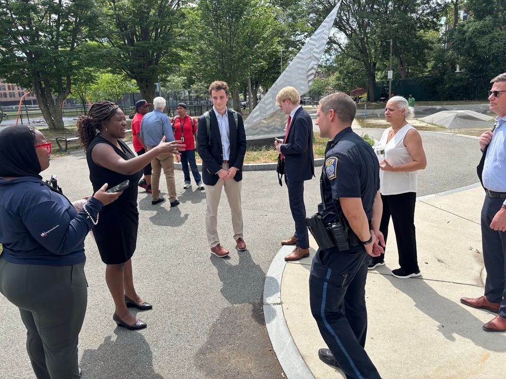 A group of people gathered around a tall white triangular monument or memorial structure. Ruthzee Louijeune in dark clothing appears to be speaking or presenting to the group, which includes men in business attire and what appears to be a uniformed officer. The setting is in a park with trees in the background.