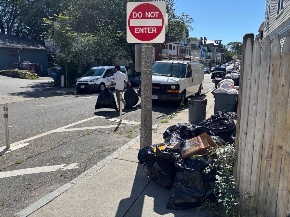 A street scene showing scattered black garbage bags and loose trash piled around a “DO NOT ENTER” sign post on a sidewalk. In the background, there are residential buildings, parked vehicles including a white van and delivery truck, and a person in white clothing. The scene appears to be on a residential street with trees and houses visible.
