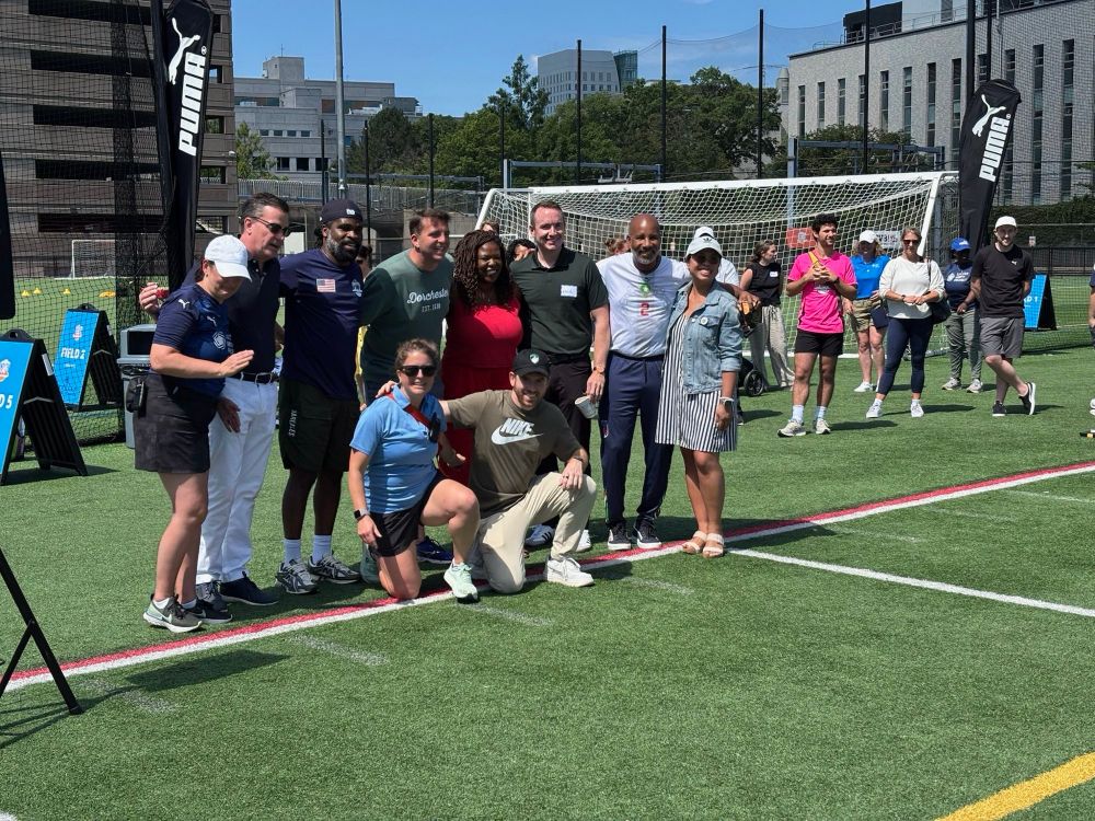 A mixed group of adults and children poses together on a soccer field with goal posts visible in the background. The group appears to be participating in a community soccer event, with participants wearing various athletic wear and casual clothing. Urban buildings can be seen beyond the field’s perimeter.​​​​​​​​​​​​​​​​