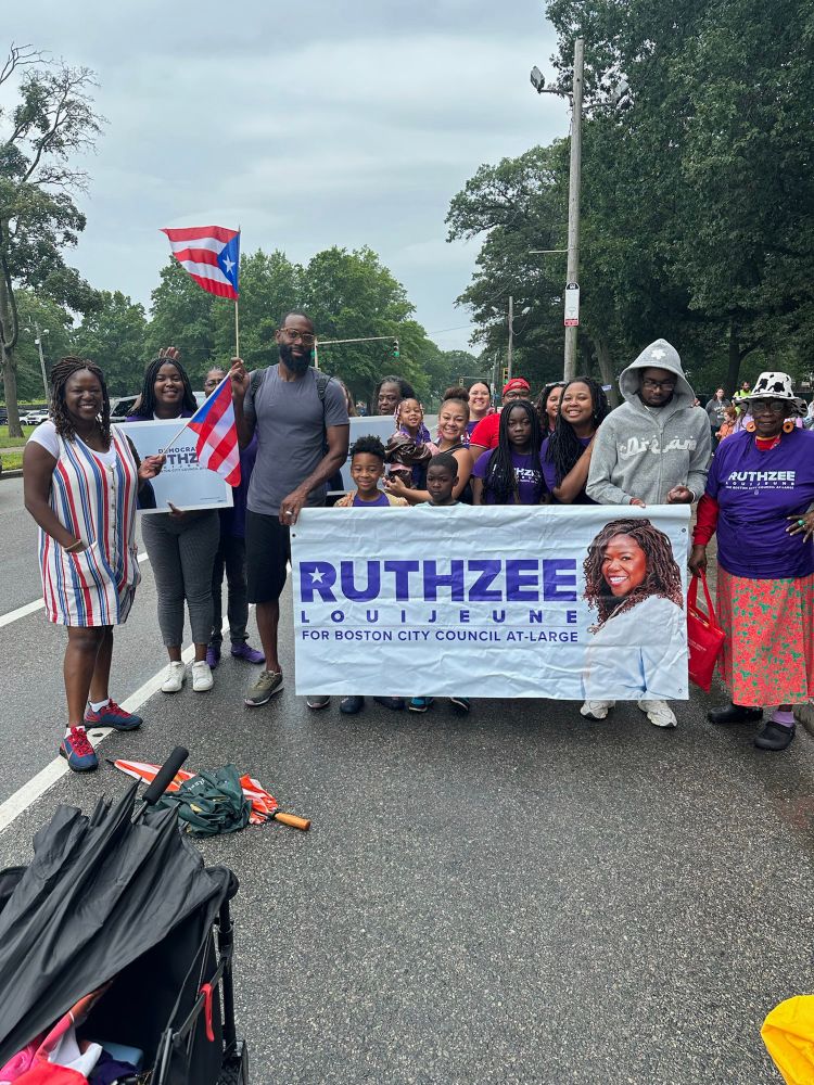 A group of campaign supporters gathered on a tree-lined street, holding a large white banner that reads “RUTHZEE LOUIJEUNE FOR BOSTON CITY COUNCIL AT-LARGE.” Ruthzee Louijeune, wearing a red, white, and blue striped dress, stands with the group. Several people are holding Puerto Rican flags, and there’s a stroller visible in the foreground. The setting appears to be during a parade or community event.