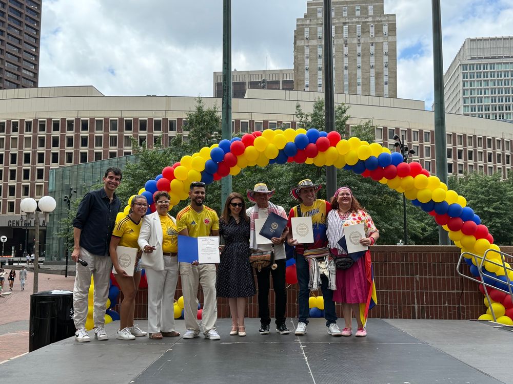  A larger group of approximately eight people pose together under an elaborate balloon arch made of red, yellow, and blue balloons at Boston City Hall Plaza. Many participants wear yellow Colombian jerseys or traditional Colombian attire, and several hold certificates or documents. The urban plaza setting features the distinctive government buildings and brick paving of City Hall Plaza.