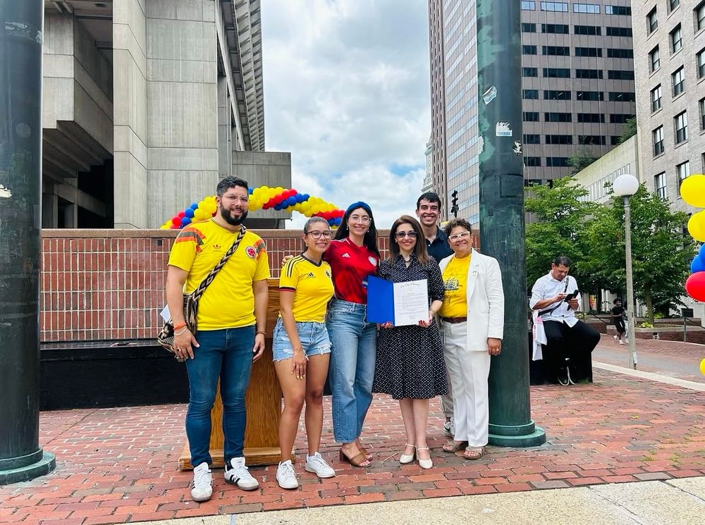 A group of seven people pose for a photo at Boston City Hall Plaza, standing in front of colorful red, yellow, and blue balloon decorations. Several people wear yellow Colombian national team jerseys, and one person holds what appears to be an official document or proclamation. The urban setting shows tall buildings and the distinctive brick plaza paving.