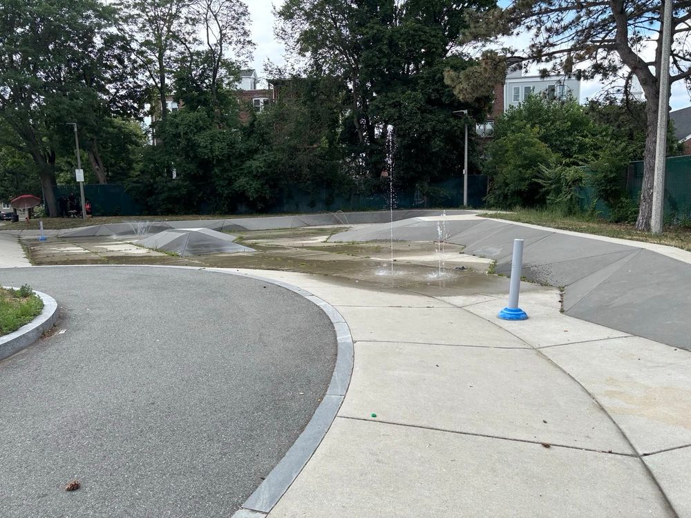 A circular concrete area with geometric raised sections and water fountains. The space appears to be a splash pad or water play area in a park setting, with trees and residential buildings visible in the background. Some of the fountain jets are active, creating water sprays.​​​​​​​​​​​​​​​​