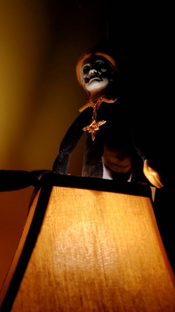 The band Ghost merch "Papa on a Pew" sitting atop of a lit lamp, the lighting and camera angle pointing upwards giving a dramatic, ominous and eerie effect to his facial features.