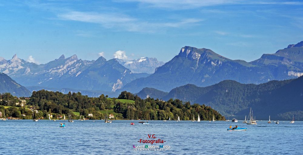 A serene lake with sailboats and kayaks under a clear blue sky. Green hills and majestic mountains form the backdrop, conveying a peaceful and vibrant atmosphere.
