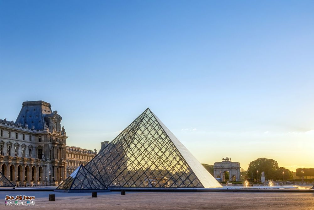Glass pyramid of the Louvre reflecting a golden sunset, framed by historic architecture and archways. Serene and majestic evening atmosphere.