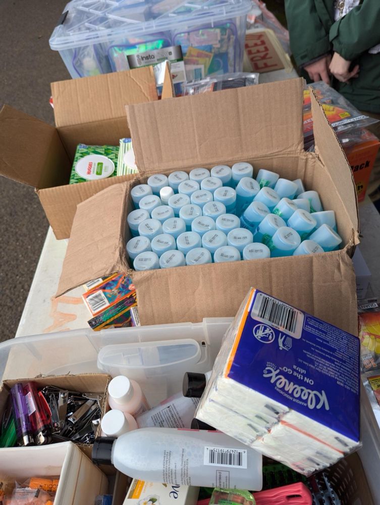 From top to bottom: a tub of packaged hygiene and bathing supplies, a box of wipes and box of hand warmers. Then a box of shampoo with Kleenex in front of it. Then a tub with clippers, cigarette lighters, sanitizer, shampoo and conditioner, soak, Kleenex, and ear plugs. 