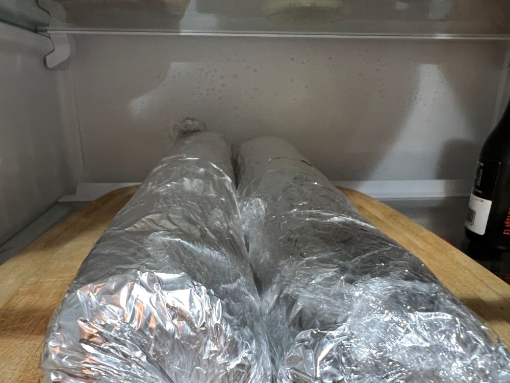 Two rolls of dough in a fridge. They're in a wooden cutting board and wrapped in foil and plastic wrap. 