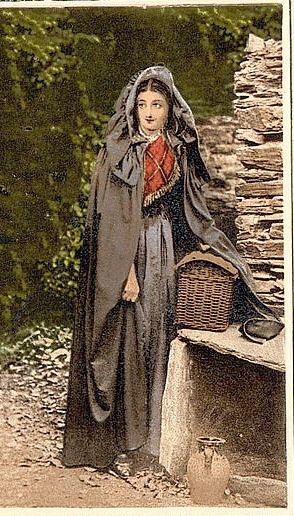 Colourised postcard image of an Irishwoman wearing a Kinsale cloak, standing next to a dry-stone wall, resting a wicker basket on a lintel. Her black cloak is floor-length, with a full, cowled hood.