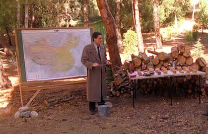 Agent Dale Cooper standing in the woods in front of a chalkboard with a map taped onto it, next to a table full of donuts in front of a big pile of firewood, with a bucket at his feet.