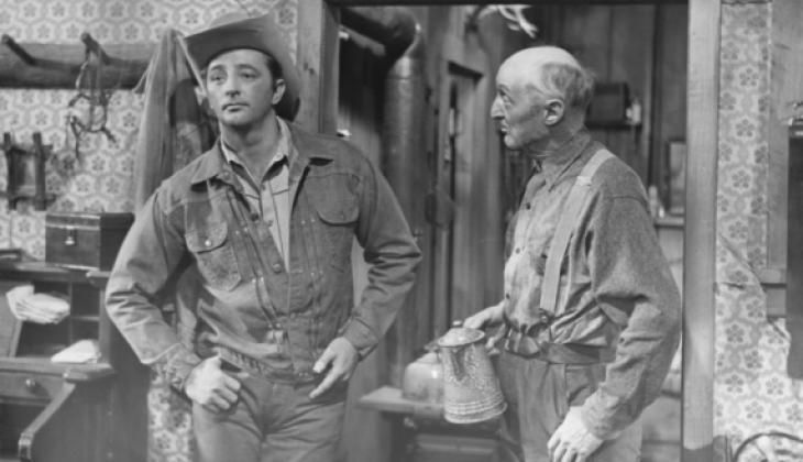 Still from a black & white western movie. Robert Mitchum talking to an older guy, leaned up against the wall looking pensively off screen and dressed as, basically, a hot cowboy. Cowboy hat, denim jacket with the top unbuttoned, hands near his hips with a thumb through his belt buckle.