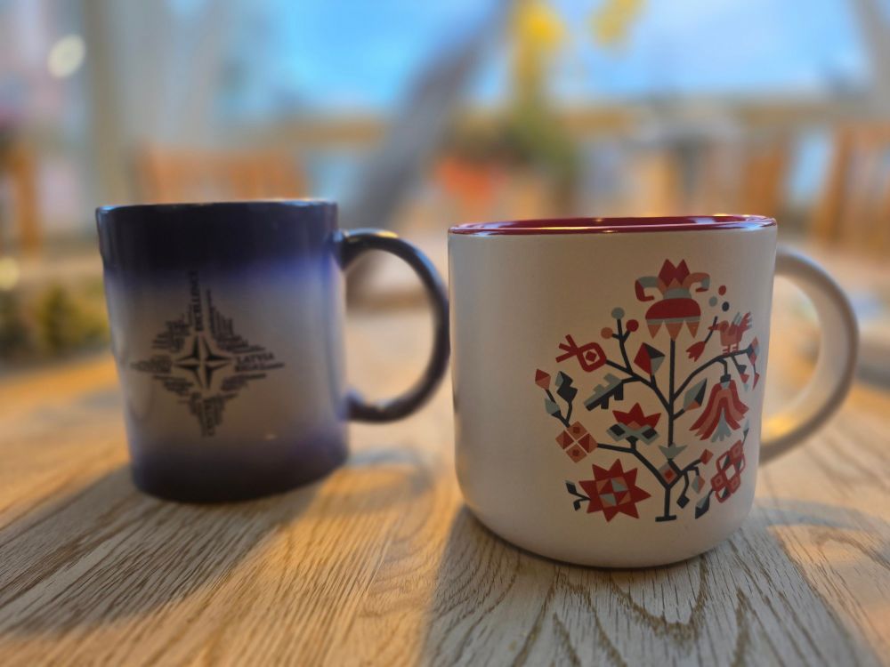 Two ceramic mugs on a wooden table with a blurred indoor background. The left mug has a gradient blue design with a NATO compass logo and text, while the right mug is white with a colorful ukrainian folk-art design featuring a tree and geometric patterns. Soft lighting creates a cozy atmosphere.

