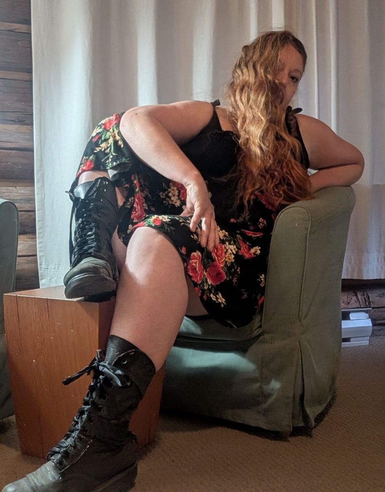 A person with long red hair is sitting on a green chair, wearing green leather boots and a rose patterned skirt. 