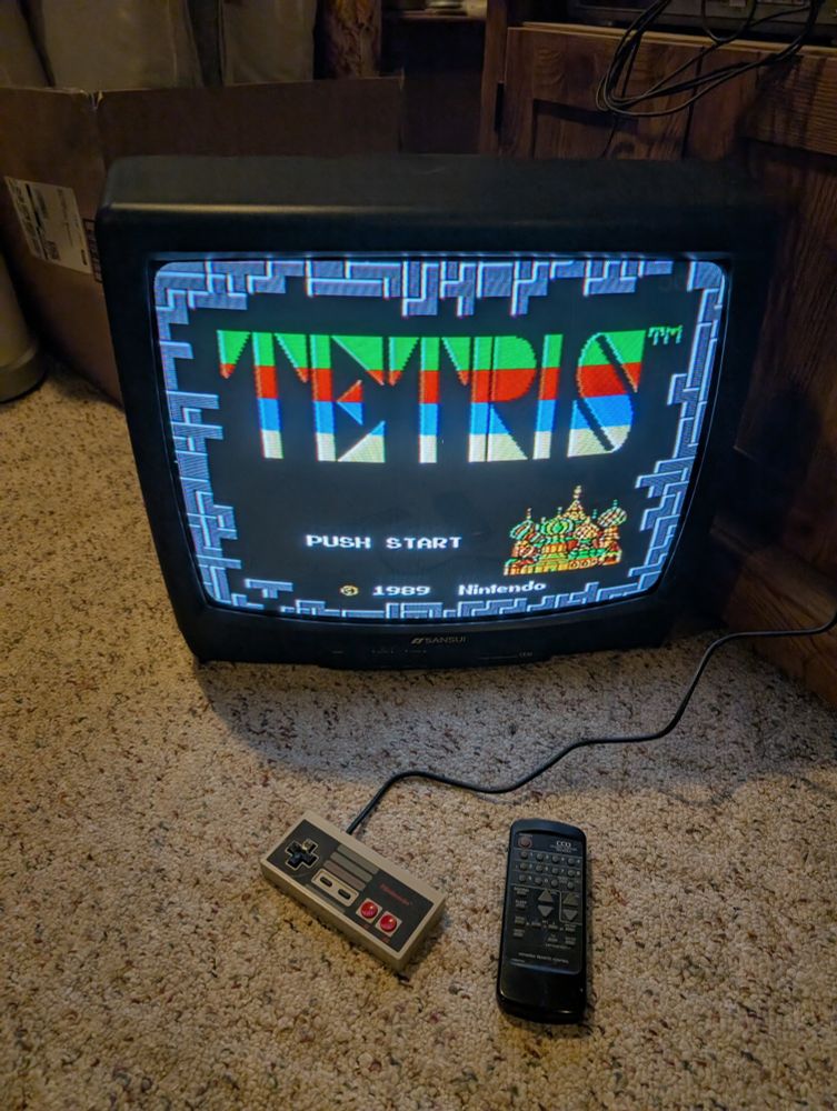 An old tube TV sits on the floor with the start screen for Tetris displayed. An NES controller and the TV remote sit in front of the TV.