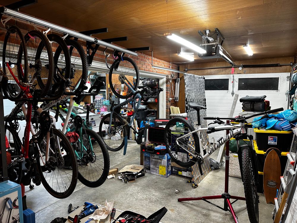 A garage with 4 bikes hanging from storage hooks and one additional bike on a work stand. The background is my filthy, not-yet-fully unpacked garage. 