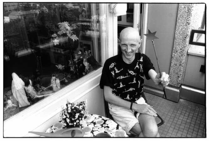 Jürgen Baldiga im August 1993 auf dem Balkon seiner Wohnung, vier Monate vor seinem Tod. Der stark von seiner AIDS-Erkrankung gezeichnete 33-Jährige hält einen langen Zauberstab (wie die gute Hexe Glinda aus dem "Zauberer von Oz") in der linken Hand und grinst den Betrachter mit leicht gebeugtem Kopf an. Auf dem runden Balkontisch Blumen und eine Gießkanne, durch das Fenster hinter ihm erkennt man unter anderem eine Marienfigur auf dem Fensterbrett im Innenraum.