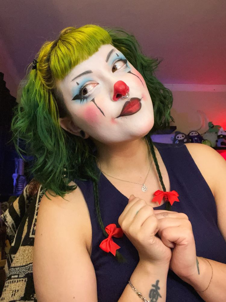 Photo of Treez in clown makeup. Their hair is green and wavy and their braids have small red bows at the ends. The makeup consists of a bright red nose and blue eyeshadow. Treez is posed innocently, looking off to the left of the photo