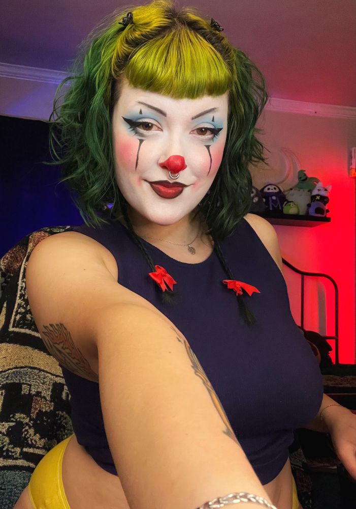Another photo of Treez in clown makeup. Their hair is green and wavy and their braids have small red bows at the ends. The makeup consists of a bright red nose and blue eyeshadow. this time their posed to look like their maybe grabbing you by the collar of your shirt. they smile sweetly regardless of their slightly more threatening energy 