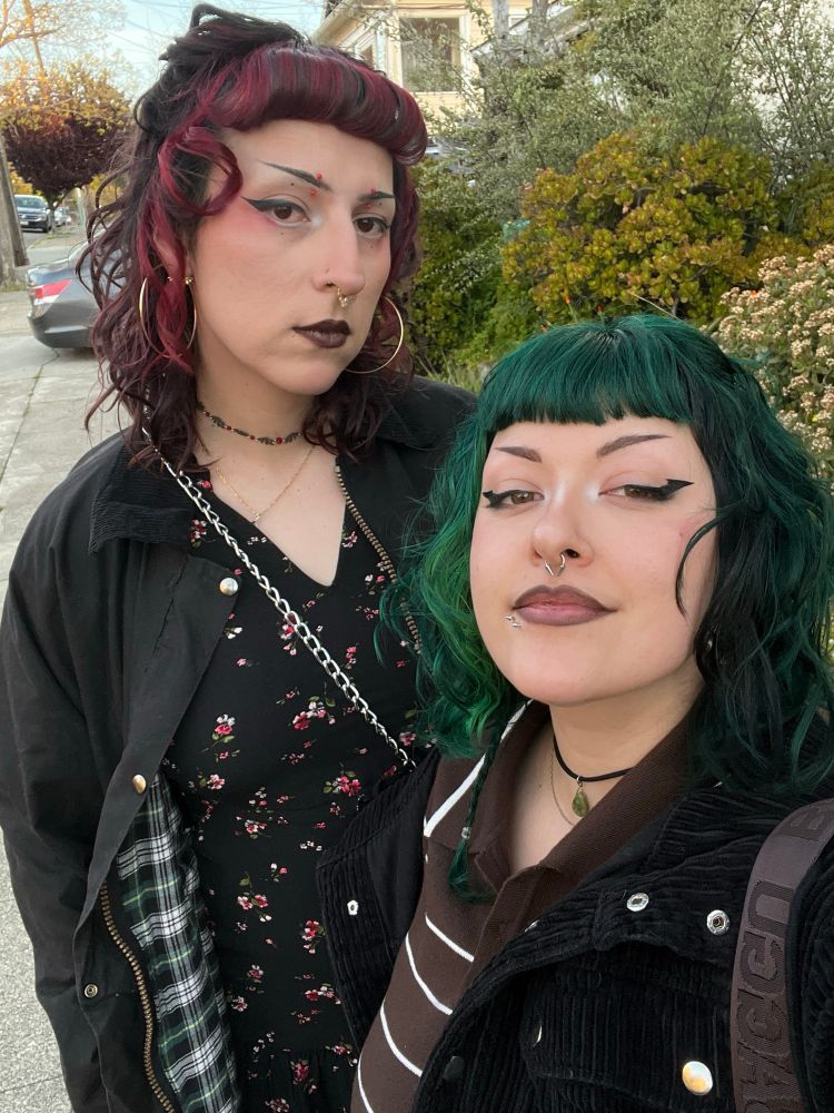 Wynter and Treez standing outside before hoping into a lyft ride. Wynter (on the left) has dark red hair with green accents and is wearing a floral dress. Treez (on the right) has short dark green hair and is wearing a stripped brown polo. they are all dressed up for their anniversary night out 