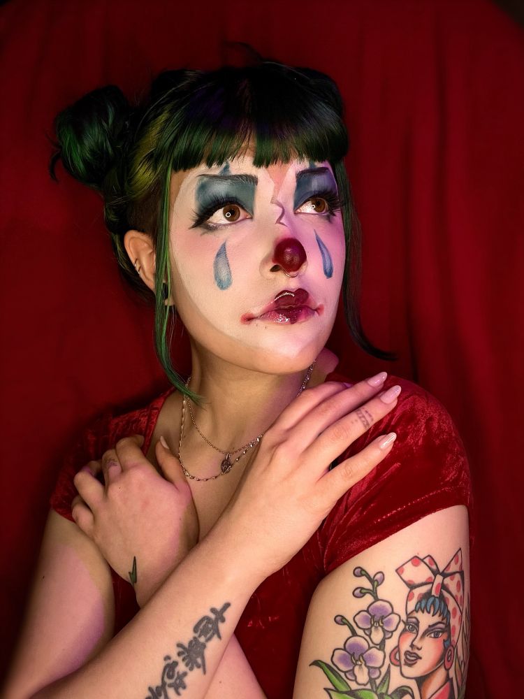 Photo of Treez from Valentine Day of 2023

Treez has their face painted like a clown inspired by an art piece that they’ve drawn in the past. The clown makeup has a broken heart white base, big blue tears on the cheeks and a heart shaped lip. Treez has their hands gently crossed cross across the chest, resting on their shoulders. Their skin is decorated in tattoos and their hair is in two buns