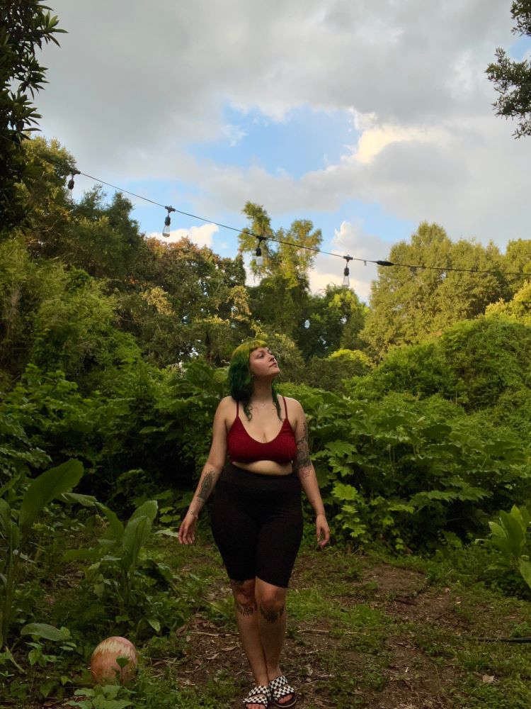 Treez, a tattooed latina with green hair, stands in an overgrown plot of land on their family’s homestead. They’re looking off into the distance, off camera. The overgrowth surrounding them is a vivid green and the setting sun illuminates the plants in a way that mirrors Treez’s hair color. The sky is full of fast moving storm clouds, breaking slightly to reveal the soft blue of the sky 