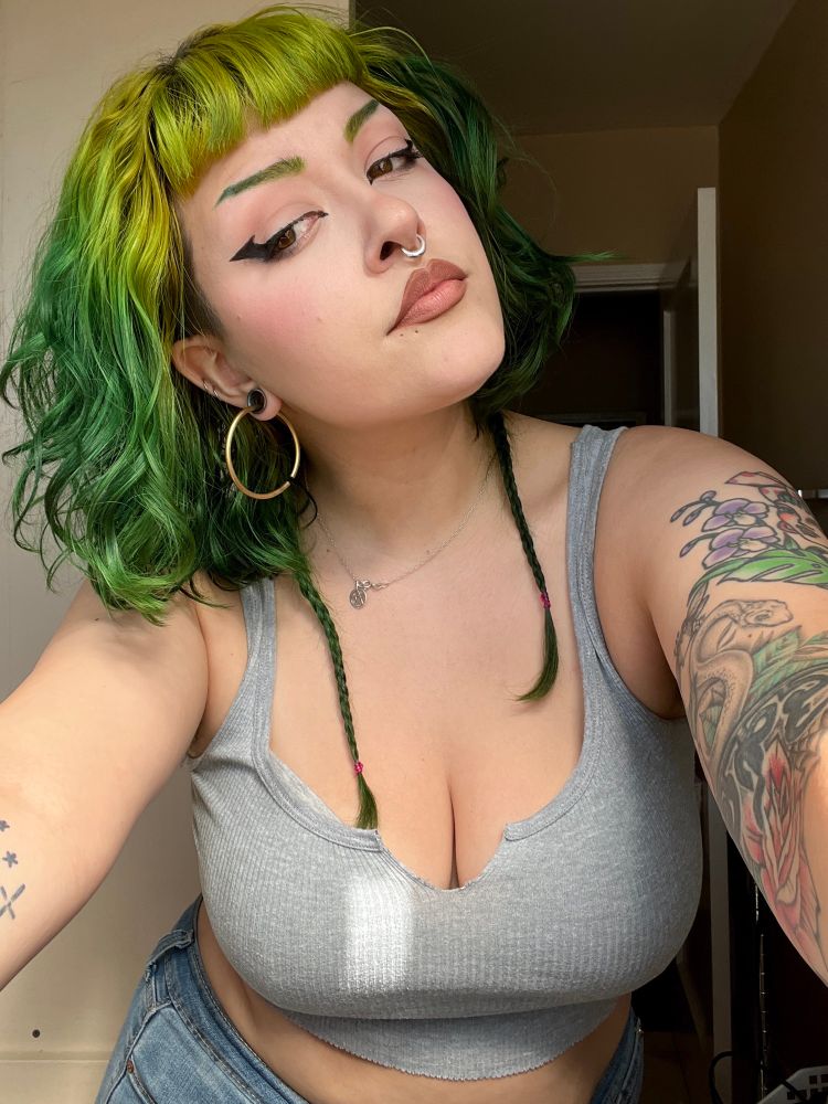 a bright sunny photo of treez, a green haired alt latina with tattoos and piercings. they are in a cropped grey tank top and are wearing jean shorts. their brows match their hair perfectly and have nice sharp eyeliner. 