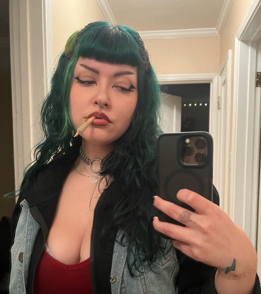 Mirror selfie of Treez: an alt Latina femme. They have long dark green hair and short bangs. They have a join in their mouth, red tank top and layered vest/zip hoodie combo. Treez has a lightning bolt tattoo on their dominant hand