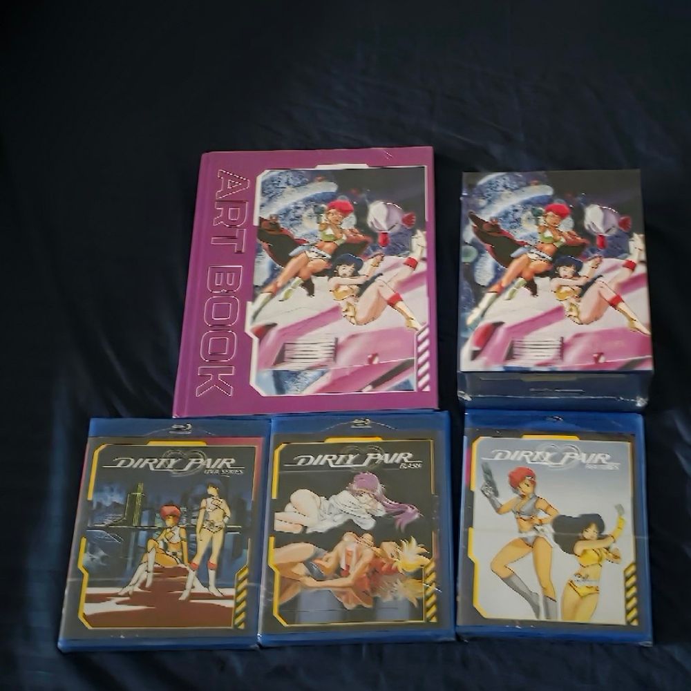 A photo taken of the bedsheet of someone’s room. On the bedsheet is an art book and several Blu-rays for the anime TV, OVA and movie series Dirty Pair. 