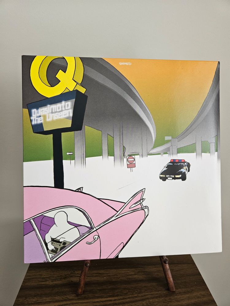 Quasimoto's debut album, 'The Unseen', on a wooden easel atop a large speaker.

The album cover features an old pink car with tail fins drives on a blank, featureless ground, underneath raised freeways, and a sign reminescent of a McDonalds sign with a large golden Q on it. Underneath the Q, it says "Quasimoto - The Unseen" A blank, featureless person in the back seat holding a boombox. A police car with its lights flashing follows the pink car.