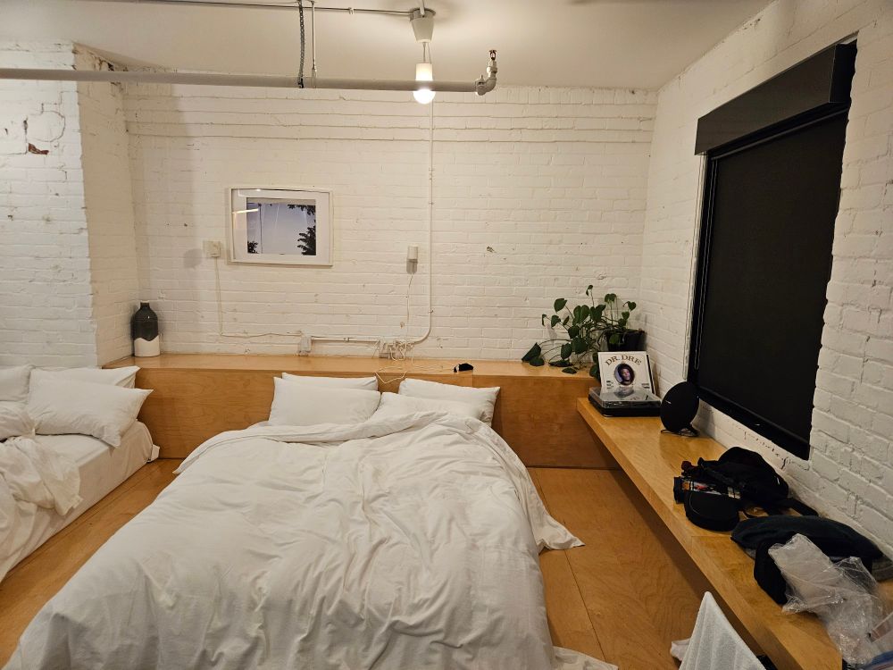 A picture of a room in The Annex, a hotel in Toronto. Includes two queen beds on a raised wooden platform, exposed brick (painted white), a live plant in the corner, and a record player with some old jazz and funk records, plus a copy of Dr Dre's first album, 'The Chronic'