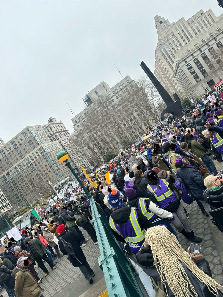 Photo of a crowd of people gathering at Foley Square. Folks from SEIU can be seen in purple vests in the foreground on the right. A green subway barrier with a yellow and green light on top divide the foreground. Thousands of people fill the center of the image going back to some large buildings in the background
