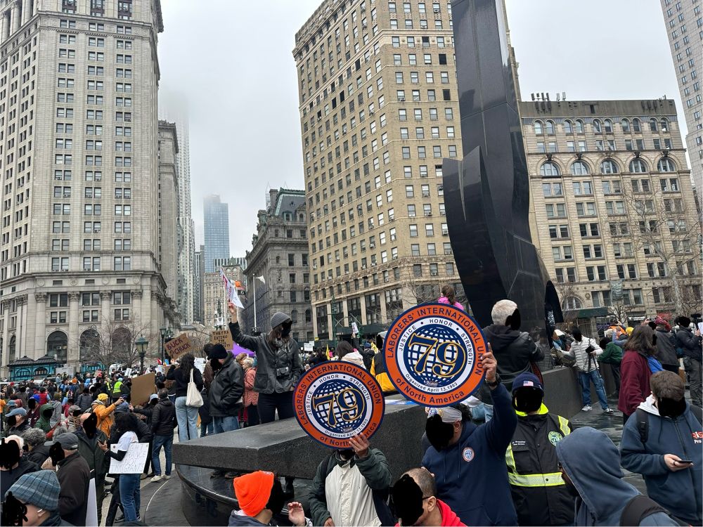 Throngs of people surround the statue at the center of Foley Square. A couple of Laborers International Union of North America 79 signs feature prominent in the middle of the foreground. Towering buildings reach into the sky. In the far distance, mist envelops the top of one particularly tall tower