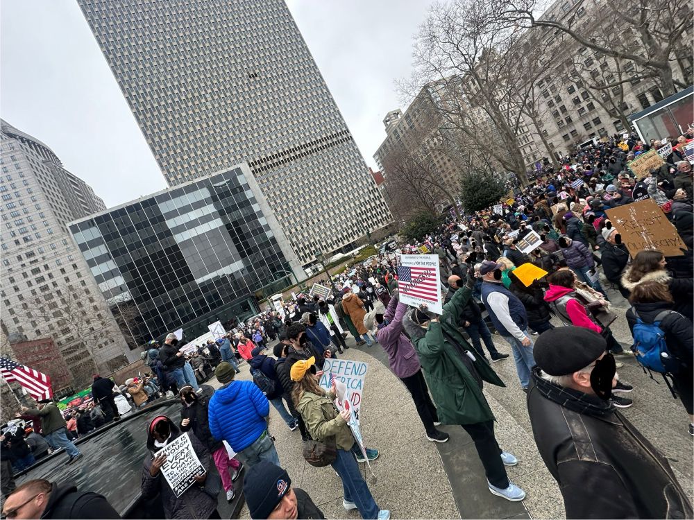 A photo from the fountain at the center of Foley Square. People hold various signs and the crowd extends to the ends of the park. Tremendous buildings reach up to the top of the photo against an overcast sky