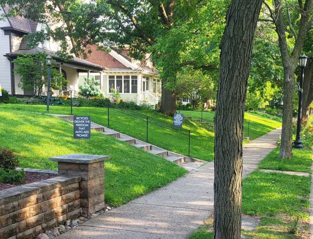 Bicycles deliver the freedom that auto adds promise.

We ask everyone outside of the car to be safe so that drivers can be dangerous. 

Two lawn signs (with the above quotes) in a sun- and shade-dappled verdant lawn gently draped before well-kept homes on a tree-lined street in a community known for its bicycle friendliness 